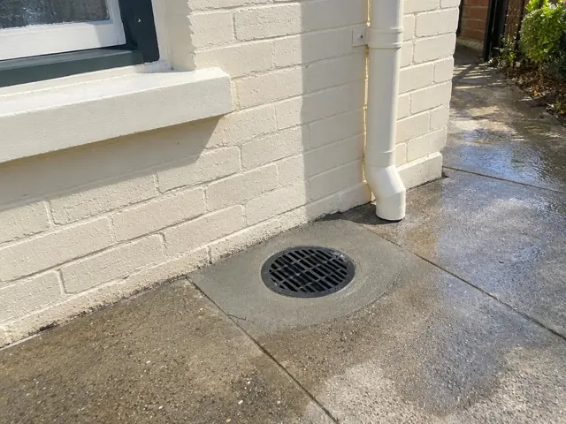 Outdoor drain grate set in concrete paving beside a cream brick house wall and downpipe in Maroubra